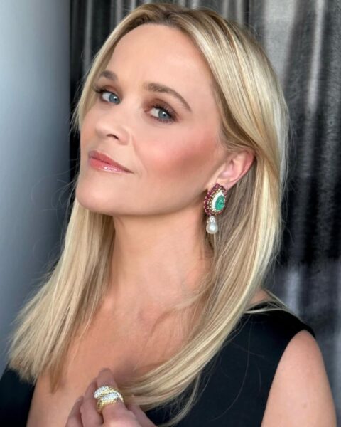 Zdjęcie aktorki Reese Witherspoon w prostych, blond włosach rozpuszczonych na ramiona. Ubrana jest w czarną sukienkę z dekoltem V. W uszach ma kolczyki z perła i białymi, czerwonymi i zielonymi kryształami. Ma wyrazisty makijaż dla dojrzałych kobiet.