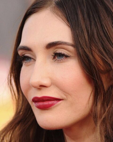 Zdjęcie aktorki Carice Van Houten ze zbliżeniem na jej makijaż. Ma gładką cerę, czerwone usta i mocno wytuszowane rzęsy. Ciemne włosy opadają jej falami po bokach twarzy.