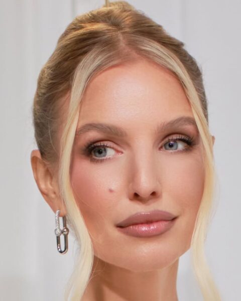 Zdjęcie kobiety z blond włosami spiętymi w kok i z kosmykami przy twarzy. Ma wiszące, srebrne, wysadzane cyrkoniami kolczyki w uszach. Ma delikatny makijaż, elegancki. Ubrana jest w koronkową, biała bluzkę. Ma też lśniącą kolię.