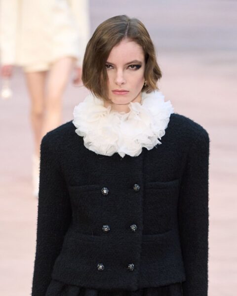 Zdjęcie modelki na wybiegu marki Dior, prezentuje modny makijaż na jesień 2025 z mocnym eyelinerem na oczach. Ma krótkie, ciemne włosy w stylu boba. Ubrana jest w czarny żakiet i ma ozdobny, biały kołnierz z falbankami przy szyi.
