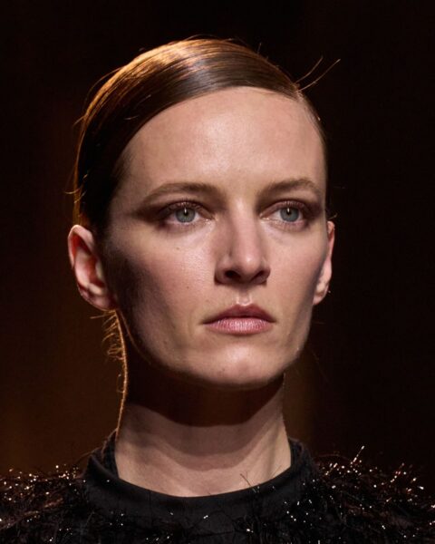 Zdjęcie portretowe modelki na wybiegu marki Elie Saab. Kobieta ma ciemne, gładko związane włosy. Ubrana jest w czarną bluzkę z cekinami. Ma wyrazisty, modny makijaż z mocno cieniowanymi oczami.