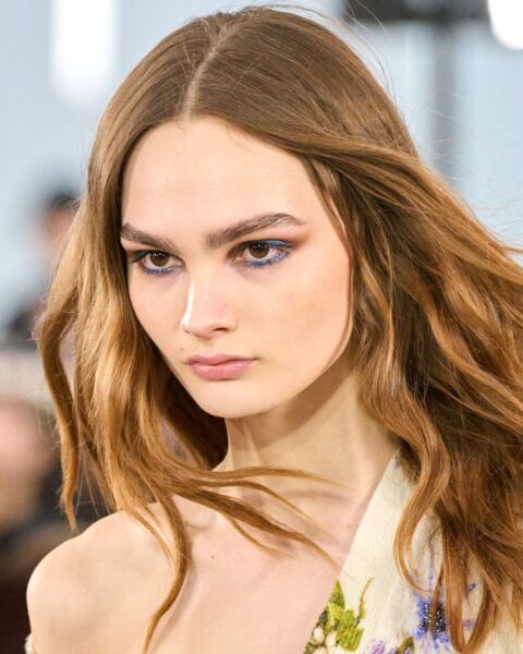 Zdjęcie portretowe modelki na wybiegu marki Blumarine. Kobieta ma ciemnoblond rozpuszczone włosy na ramiona. Ubrana jest w asymetryczną sukienkę w kolorze beżowym z nadrukiem kwiatów, odsłaniającą jedno ramię. Ma wyraziście pomalowane oczy w kolorze brązu i niebieskości.