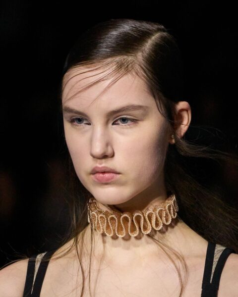 Zdjęcie portretowe modelki na wybiegu marki Dior. Ma ciemne, rozpuszczone włosy. Ubrana jest w czarny top na ramiączkach. Na szyi ma masywny, złoty naszyjnik z kształcie fal. Ma oryginalnie zaaplikowany eyeliner w kącikach wewnętrznych oczu.