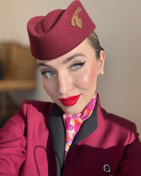 Zdjęcie stewardessy robiącej sobie selfie. Ma na głowie bordową czapkę, a na sobie bordowy uniform z czarnym kołnierzem. Na szyi ma kolorową apaszkę. Makijaż z wyraźnymi ustami i czarną kreską eyelinerem przy rzęsach. Ma gładko związane blond włosy.