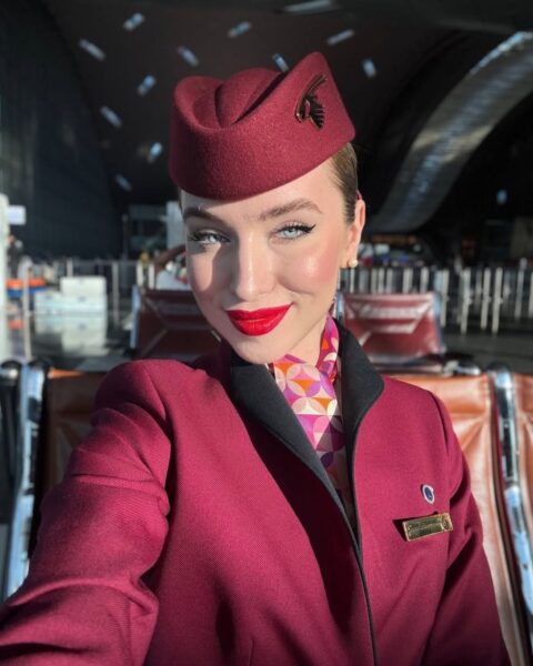 Zdjęcie kobiety na lotnisku, która zrobiła sobie selfie. Prezentuje makijaż stewardessy z różowymi policzkami, kreską na powiece i czerwonymi ustami. Ma na sobie bordową czapkę lotniczą, uniform oraz ozdobną chustę.