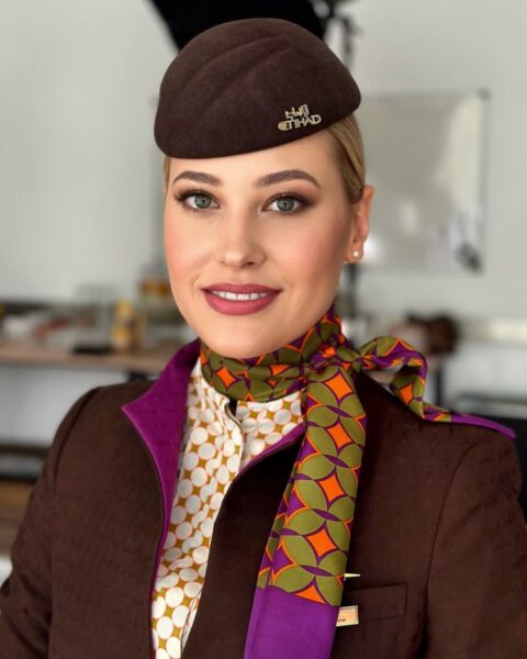 Zdjęcie stewardessy. Ma delikatny, upiększający makijaż. Na głowie czapkę brązową, brązowy uniform z fioletową podszewką, koszulę w biało-zielono-pomarańczowy wzór oraz apaszkę w zielono-fioletowo-pomarańczowy wzór. Ma blond włosy, gładko spięte.