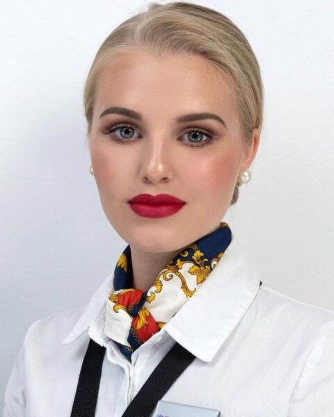 Zdjęcie makijażu stewardessy. Ma delikatnie pomalowane oczy, świetlistą cerę i czerwone usta. Włosy blond, spięte gładko. Ubrana jest w biała koszulę, a na szyi ma wzorzystą koszulę. W uszach perłowe kolczyki.
