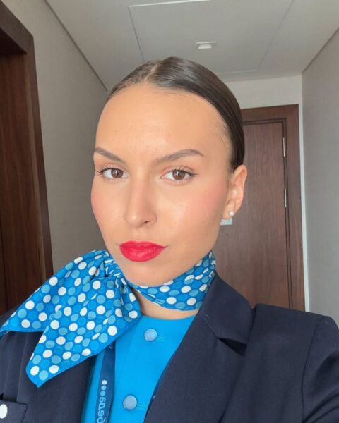 Zdjęcie kobiety, która pokazuje swój profesjonalny makijaż stewardessy, czyli gładką cerę i czerwone usta. Jest ubrana w niebieski uniform, granatową marynarkę, a na szyi ma niebieską apaszkę. Włosy ciemne, gładko związane.