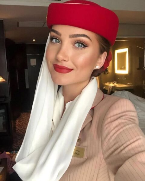 Zdjęcie stewardessy, która zrobiła sobie selfie. Ma wyrazisty makijaż oczu z cieniami, eyelinerem i mocno wytuszowanymi rzęsami. Ubrana jest w beżowy, prążkowany uniform, a na głowie ma czerwoną czapkę oraz fragment chusty jako znak rozpoznawczy linii arabskich.