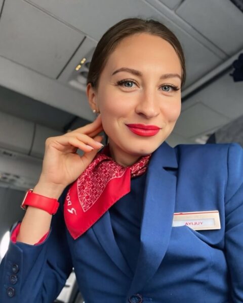 Zdjęcie stewardessy robiącej sobie selfie na pokładzie. Siedzi na fotelu, podpiera ręką głowę. Ma czerwony zegarek. Ubrana jest w granatowy uniform i ma na szyi czerwoną apaszkę. Włosy blond, związane z tyłu. Ma czerwone usta i wytuszowane rzęsy.