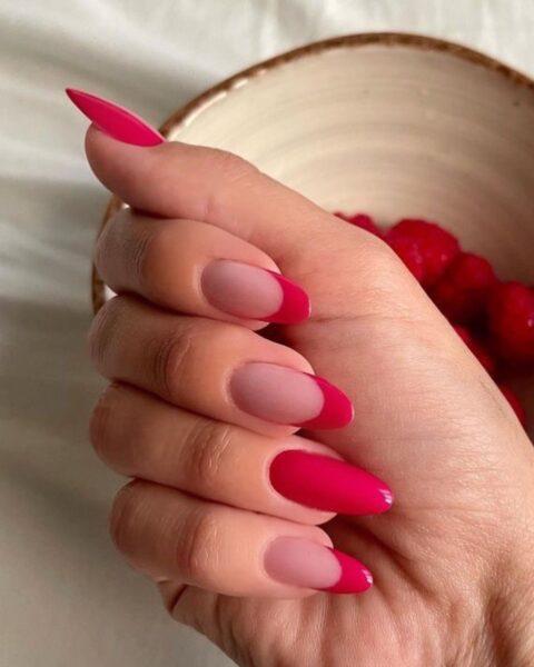 Zdjęcie dłoni z długimi paznokciami o migdałowym kształcie ozdobione wzorem french manicure w malinowym kolorze. Dłoń pozuje nad miseczką z malinami.