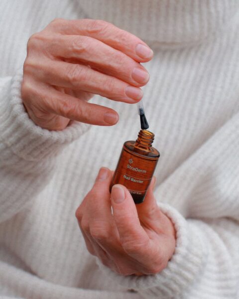 Zdjęcie przedstawiające manicure dla 60-latki. Ciasny kadr skoncentrowany na dłoniach seniorki, która maluje paznokcie odżywką. Dłonie pozują na tle beżowego swetra.