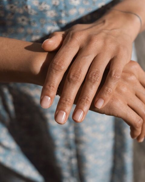 Zdjęcie dłoni ze zbliżeniem na krótkie, owalne paznokcie pokryte odżywką. Dłonie układają się tak, by prezentowac manicure. Dłonie pozują na tle niebieskiej, kwiecistej sukienki.