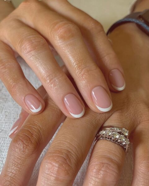 Zdjęcie dłoni z długimi, owalnymi paznokciami. To french manicure dla 60-latki. Widać na palcu serdecznym dwa srebrne pierścionki z brylantami. Dłonie pozuje na tle białego ręcznika.