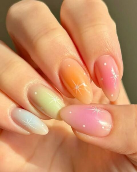 Zdjęcie dłoni z długimi, migdałowymi paznokciami z manicure summer aura nails, czyli w pastelowym kolorze, każdy paznokieć ma inny kolor. Do tego jest ozdobiony gwiazdkami, kropkami i kryształkami. Dłoń pozuje tak, by prezentować manicure.