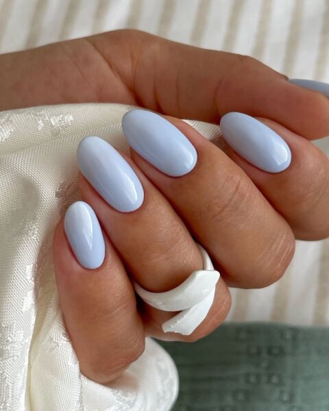 Zdjęcie dłoni z długimi, migdałowymi paznokciami. Są pomalowane na kolor jasnego błękitu w odcieniu baby blue. Dłoń trzyma wyszywany, kremowy materiał. Na środkowym placu znajduje się biały pierścionek.
