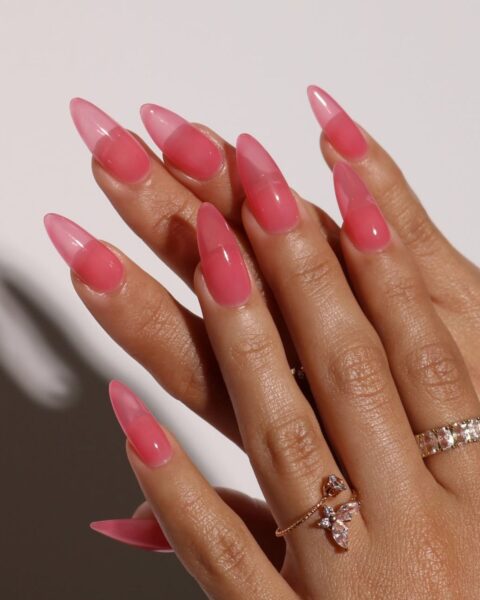 Zdjęcie dłoni z długimi, migdałowymi paznokciami z manicure w stylu jelly nails w kolorze koralowym. Na palcach znajduje się kilka pierścionków. Dłonie pozują tak, by prezentować paznokcie.