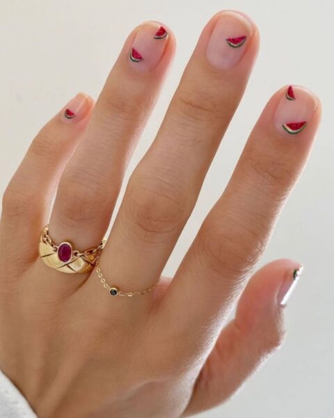 Zdjęcie dłoni z krótkimi, owalnymi paznokciami w kolorze nude, ozdobione wzorem micro fruit manicure z motywem małych arbuzów. Na palcach znajduje się kilka złotych pierścionków.