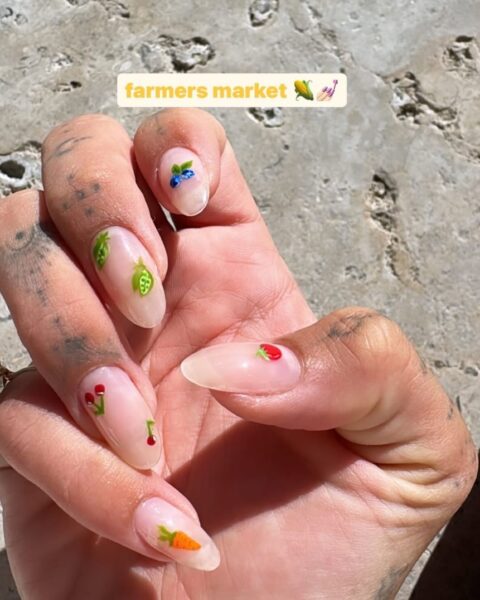 Zdjęcie dłoni ze zbliżeniem na micro fruit manicure na długich paznokciach. Są to paznokcie celebrytki Hailey Bieber. Ma na palcach tatuaże.