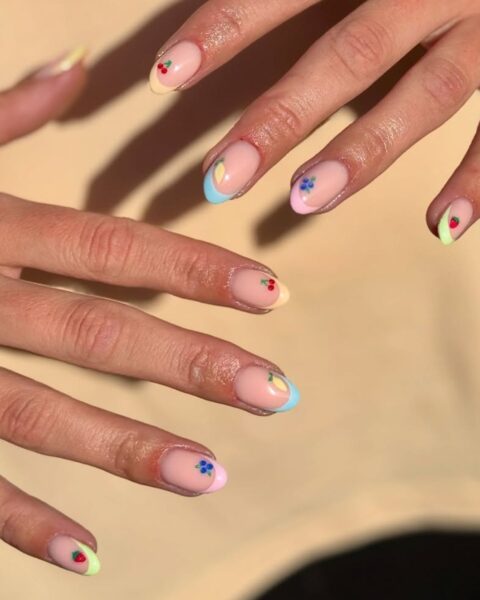 Zdjęcie dłoni z długimi, owalnymi paznokciami pomalowanymi na kolorowy manicure z różnobarwnymi końcówkami oraz ozdobione wzorem drobnych owoców. Dłonie pozują na tle żółtej koszulki.