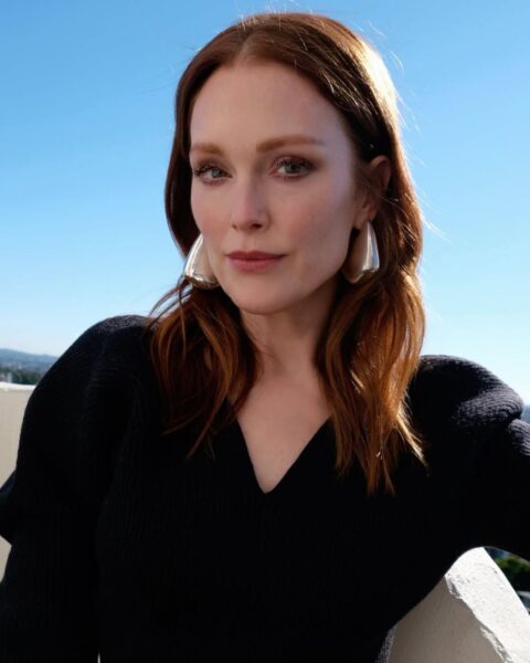 Zdjęcie aktorki Julianne Moore, która ma modne włosy na lato 2025, czyli falowane, długie do ramion i w kolorze rudym. Ubrana jest w czarny sweter z bufkami. W uszach ma srebrne kolczyki.