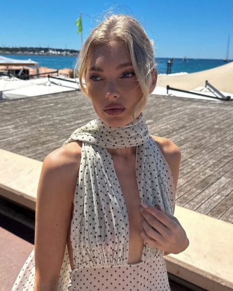 Zdjęcie modelki i influencerki Elsy Hosk. Ma jasnoblond włosy w odcieniu scandi blonde spięte w kok z kosmykami przy twarzy. Ubrana jest w zwiewną sukienkę bez rękawów i wiązaniem wokół szyi. Sukienka jest kremowa i w czarne groszki. Pozuje w ciepłym kurorcie.
