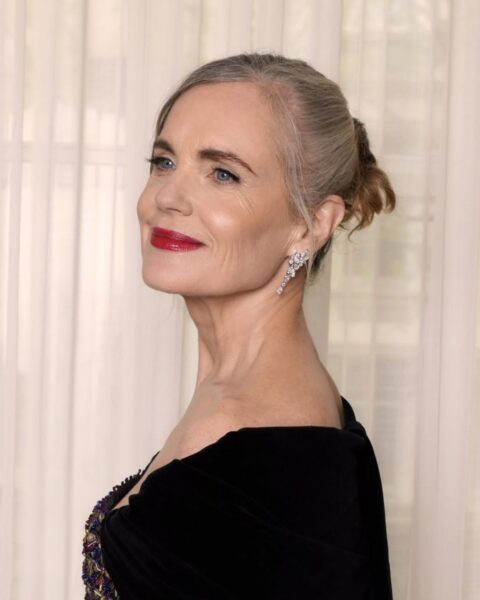 Zdjęcie aktorki Elizabeth McGovern, która ma naturalnie siwe włosy. Na zdjęciu ma je związane z tyłu i ma kosmyki przy twarzy. Ubrana jest w wieczorową suknię z szerokim kołnierzem odkrywającym ramiona. W uszach ma kolczyki z brylantami. Ma wieczorowy makijaż z czerwonymi ustami. Pozuje na tle okna z białą zasłoną, patrząc w dal.