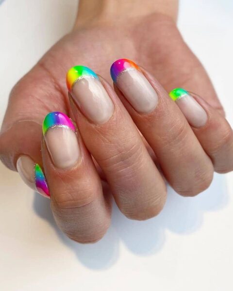 Zdjęcie dłoni z długimi, owalnymi paznokciami pomalowanymi w stylu french manicure w tęczowym ombre. Dłoń pozuje na białym blacie i prezentuje manicure.
