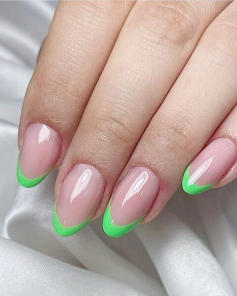Zdjęcie dłoni z długimi, owalnymi paznokciami pomalowanymi w stylu french manicure z neonową, zieloną końcówką. Dłoń pozuje na tle białego materiału z satyny.