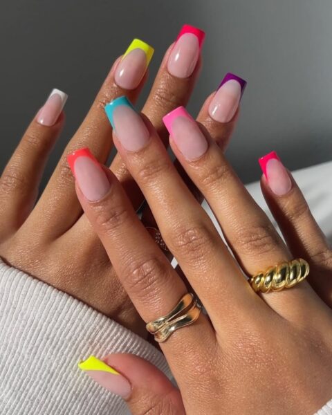 Zdjęcie dłoni ze zbliżeniem na neonowy french manicure na lipiec: widać kwadratowe dłonie z kwadratowymi paznokciami. Każdy paznokieć ma inny kolor. Na palcach znajdują się złote pierścionki.