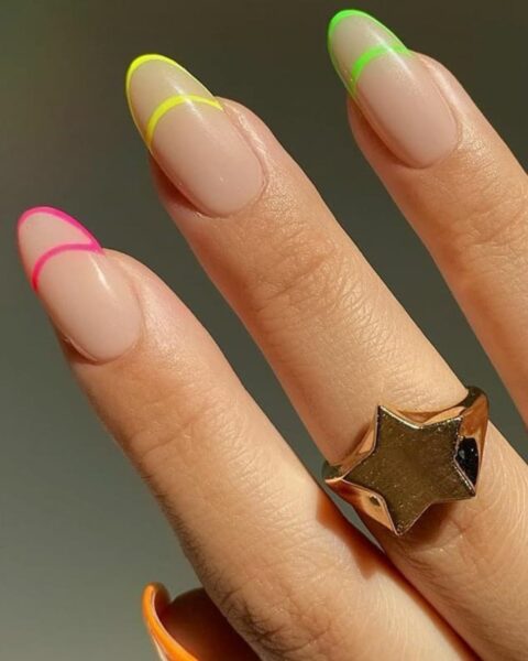 Zdjęcie dłoni ze zbliżeniem na długie, owalne paznokcie ozdobione wzorem double french manicure w neonowych kolorach. Na każdym paznokciu jest inny kolor. Na palcu widać złoty pierścionek z motywem gwiazdy.