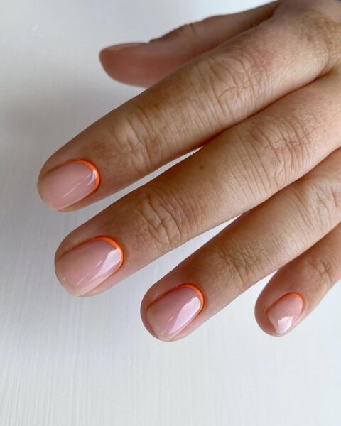 Zdjęcie dłoni z krótkimi, owalnymi paznokciami ozdobione wzorem reverse french manicure w kolorze pomarańczowym w formie paska u nasady paznokci.
