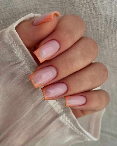 Zdjęcie dłoni z długimi, kwadratowymi paznokciami. Zdobi je neonowy french manicure na lipiec 2025 w kolorze brzoskwiniowym. Dłoń pozuje w lnianym, jasnym rękawie bluzki.