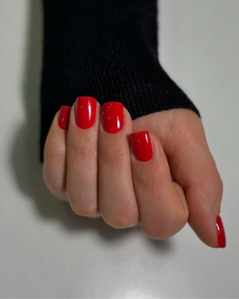 Zdjęcie dłoni z krótkimi, kwadratowymi paznokciami w kolorze czerwonym. Dłoń pozuje na tle białego blatu prezentując manicure.