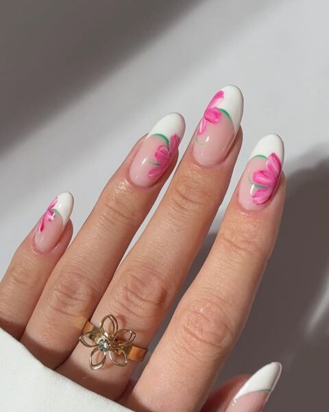Zdjęcie dłoni ze zbliżeniem na długie, owalne paznokcie. Są pomalowane w stylu french manicure i ozdobione wzorem kwiatów wykonanych techniką nail artu. Na palcu środkowym widać złoty pierścionek z motywem kwiatka.