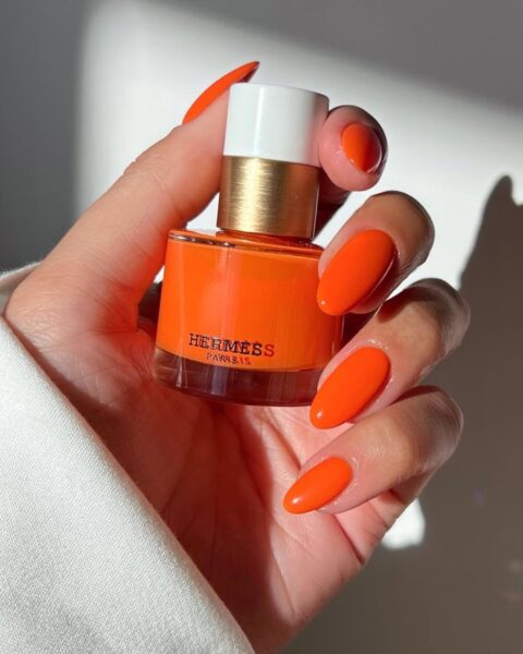 Zdjęcie dłoni ze zbliżeniem na długie, migdałowe paznokcie pomalowane na kolor pomarańczowy w odcieniu tangerine. Dłoń trzyma buteleczkę lakieru do paznokci Hermes.