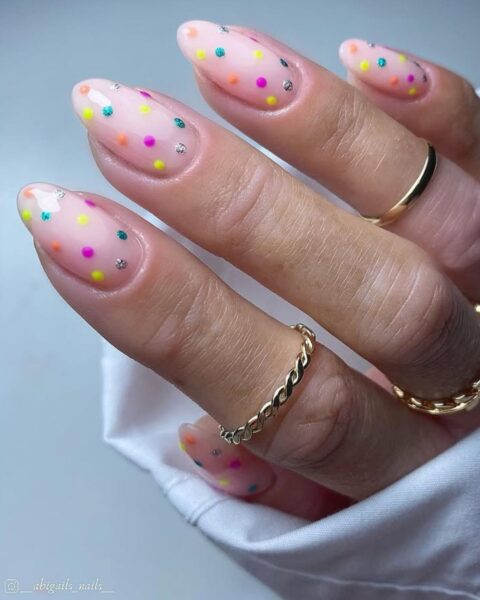 Zdjęcie dłoni z długimi, migdałowymi paznokciami w kolorze nude, ozdobione wzorem polka dots w tęczowych barwach. ?na palcach znajduje się kilka złotych paznokci.