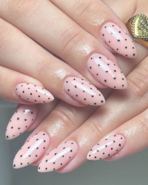 Zdjęcie dłoni z długimi, migdałowymi paznokciami. Są pomalowane wzorem polka dots nails, czyli w kolorze nude i w czarnymi groszkami. Dłonie układają się tak, by prezentować manicure.