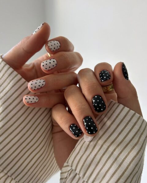 Zdjęcie dłoni ze zbliżeniem na krótkie, owalne paznokcie polka dots. Są pomalowane na kolor biały i czarne groszki na jednej dłoni, a na drugiej czarne z białymi groszkami. Widać liczne złote pierścionki oraz rękawy koszuli kremowej w oliwkowe prążki.