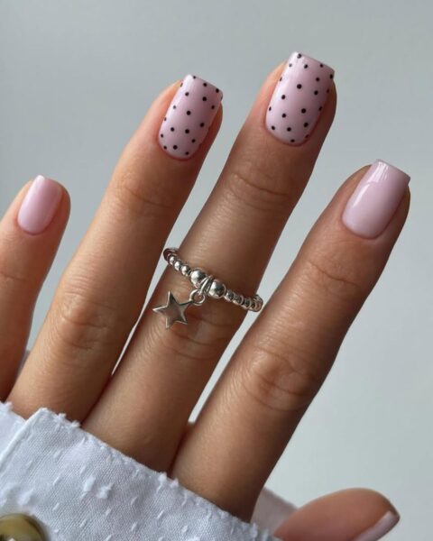 Zdjęcie dłoni ze zbliżeniem na długie, kwadratowe paznokcie w kolorze pink nude i ozdobione w stylu polka dots manicure, czyli na dwóch paznokciach są czarne, drobne groszki. Na środkowym palcu widać srebrny pierścionek z motywem gwiazdki.