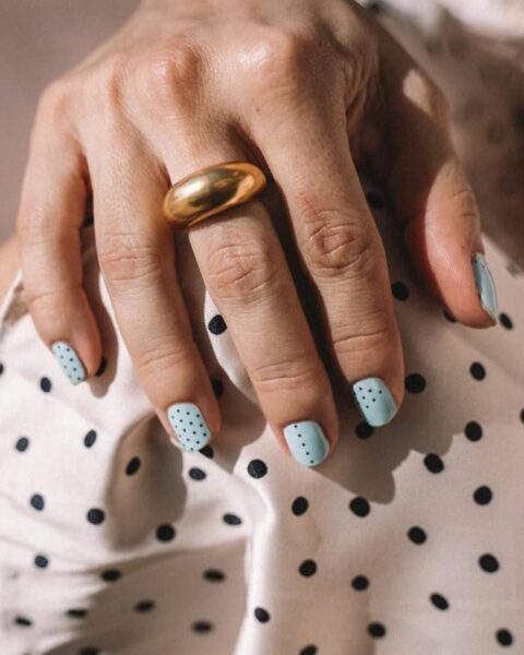 Zdjęcie dłoni ze zbliżeniem na paznokcie polka dots. Są krótkie, owalne, pomalowane na biało i w czarne groszki. Na palcu widać masywny, złoty pierścionek. Dłoń opiera się o ramię. Widać bluzkę białą w czarne groszki.