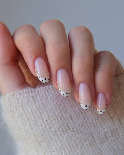 Zdjęcie dłoni z długimi, migdałowymi paznokciami. Są pomalowane na wzór french manicure, gdzie biała kreska na brzegu jest ozdobiona czarnymi kropkami.