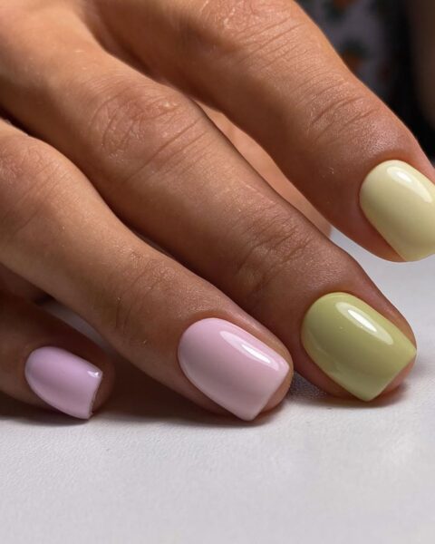 Zdjęcie dłoni ze zbliżeniem na kwadratowe paznokcie w kolorze fioletowym i zielonym. Dłoń opiera się o biały blat stołu i pozuje tak, by prezentować manicure.