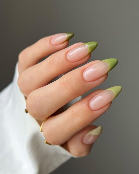 Zdjęcie dłoni ze zbliżeniem na długie, migdałowe paznokcie ze stylizacją french manicure, ale zamiast białych końcówek są zielone. Widać na palcach złote pierścionki oraz fragment białego rękawa bluzki.