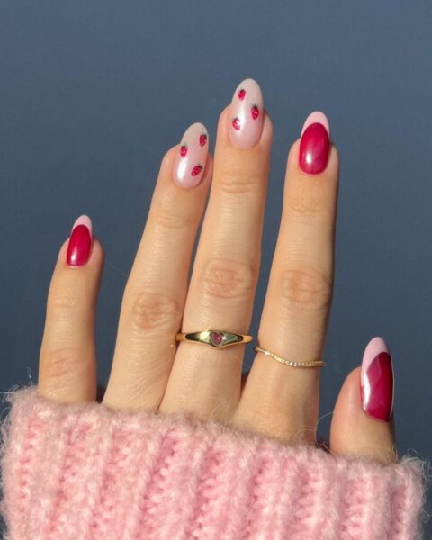 Zdjęcie dłoni z długimi, owalnymi paznokciami. Są pomalowane na wzór french manicure z jasnym i ciemnym różem. Dwa paznokcie są pomalowane na jasny róż i ozdobione wzorem poziomek. Na palcach znajdują się dwa pierścionki.
