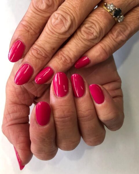 Zdjęcie dojrzałych dłoni, które prezentują manicure w kolorze poziomkowego różu. Na palcu serdecznym znajduje się złoty pierścionek z brylantem. Dłonie układają się tak, by prezentować manicure.
