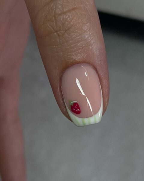 Zdjęcie kciuka ze zbliżeniem na paznokieć z french manicure z końcówką w biało-zielone paski. Do tego pojawia się wzór drobnej poziomki.