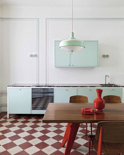 Zdjęcie dużej kuchni o minimalistycznym designie. Widać szafki w miętowym kolorze, wiszącą lampę w kolorze miętowym, a na pierwszym planei drewniany stól z czzrwonymi nogami i czerwonymi dekoracjami na jego blacie. Wokół niego stoją drewniane krzesła na czerwonych nogach. Z kolei podłoga wyłożóna jest kafelkami w kolorze kremowo-czerwonym układającymi się we wzór szachownicy.