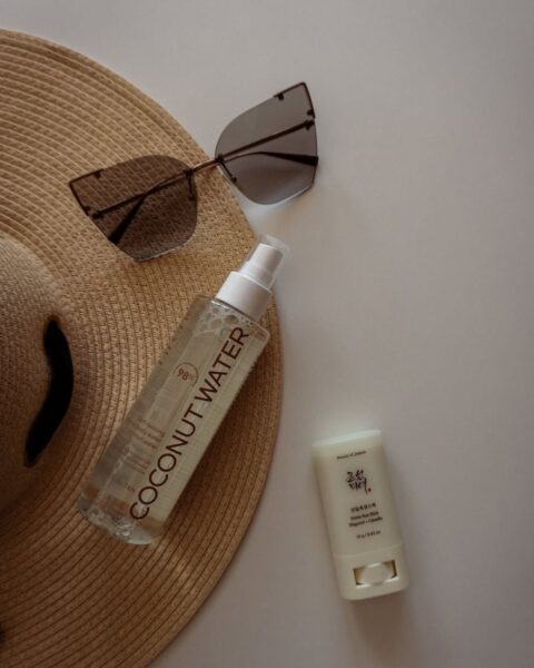Zdjęcie przedstawiające rodzaje filtrów SPF: mgiełkę kokosową oraz krem SPF w sticku. Jest to zdjęcie typu flat lay, widać od góry kosmetyki, a do tego książkę, słomkowy kapelusz i okulary przeciwsłoneczne.