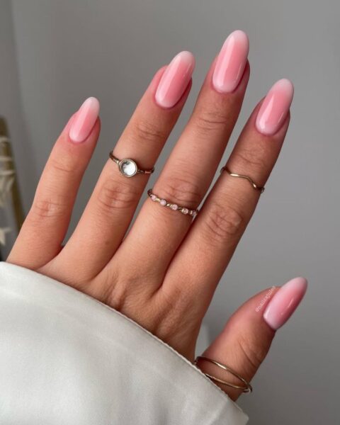 Zdjęcie dłoni ze zbliżeniem na długie, owalne paznokcie z efektem blushed nails w stylu scandi-chic manicure. Na palcach znajdują się pierścionki. Widać też fragment białego rękawa.